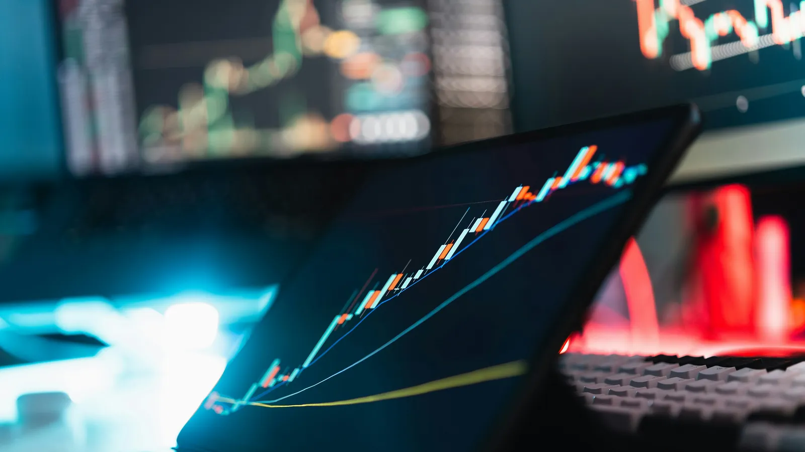 Tablet showing a rising candlestick chart in a dark room, with blurred dashboards glowing behind it