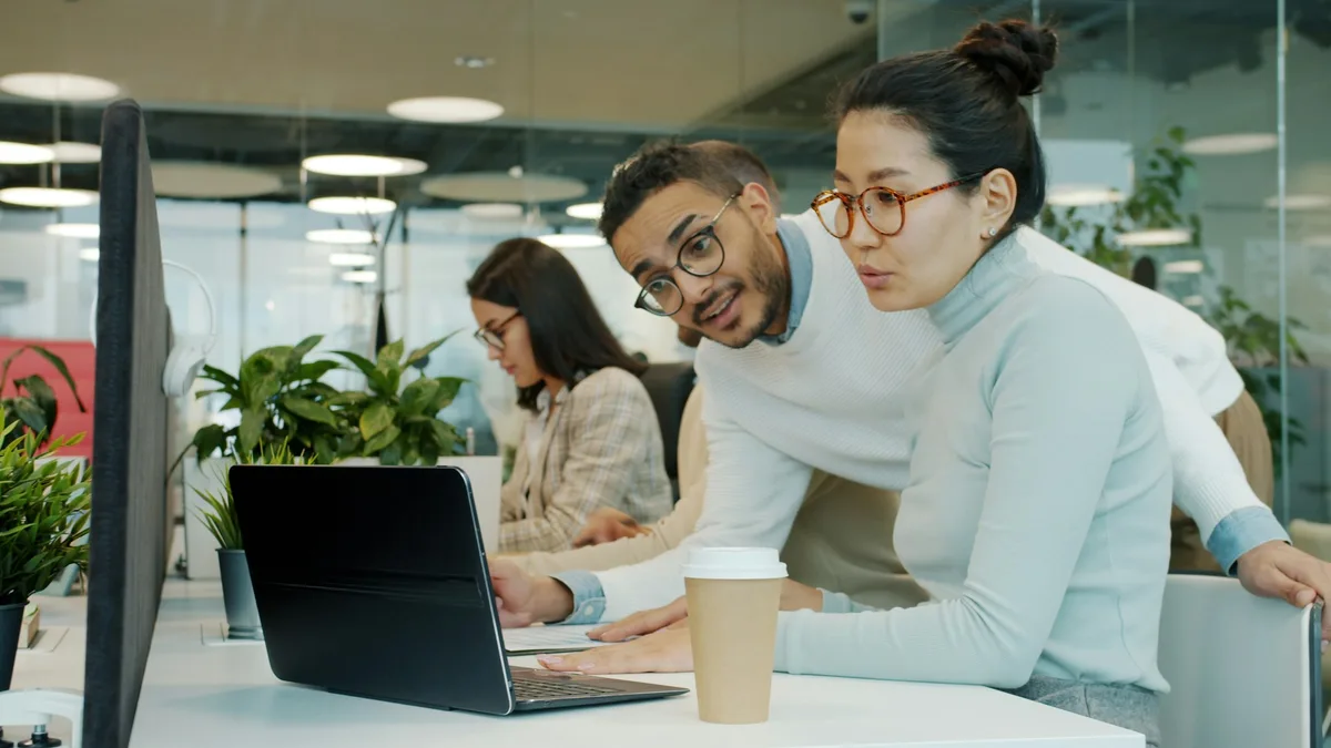 Two colleagues collaborating and discussing work on a laptop in an office