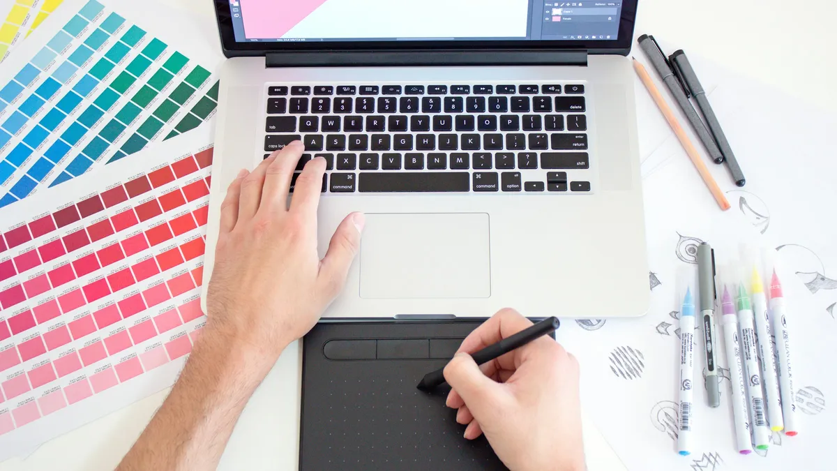 Designer working across a tablet and laptop with printed color swatches on the desk