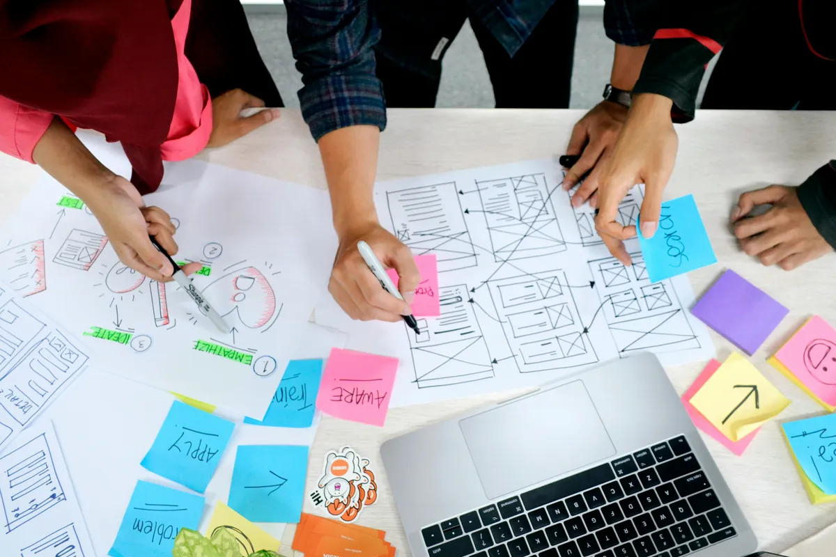 Designers sketching UI wireframes and hierarchy on paper with sticky notes and a laptop