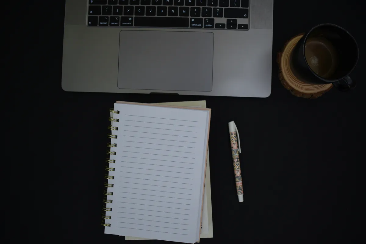 An open lined notebook and pen beside a laptop on a dark desk ready for a handoff note