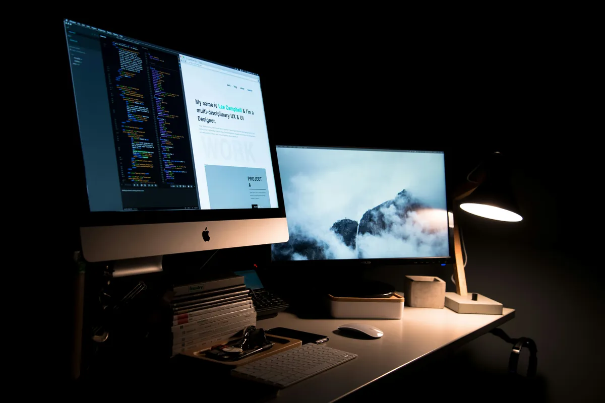 Silver iMac and a second monitor glowing on a dark desk with code open on the left screen