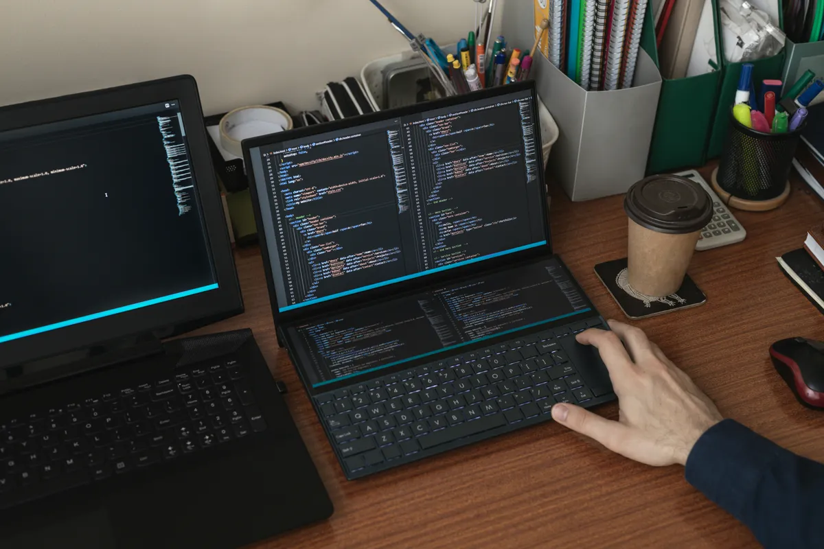 Hand on the trackpad of a laptop showing code, a second coding laptop beside it