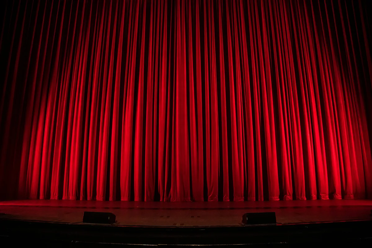 A red velvet stage curtain hanging closed above an empty stage with two small footlights