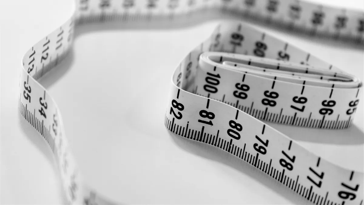 Black and white close-up of a cloth tape measure curling across a pale surface, marks 76 to 100